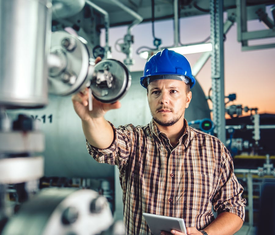 Male engineer wearing a blue hard hat inspecting industrial machinery and using a tablet for equipment monitoring in a manufacturing facility.