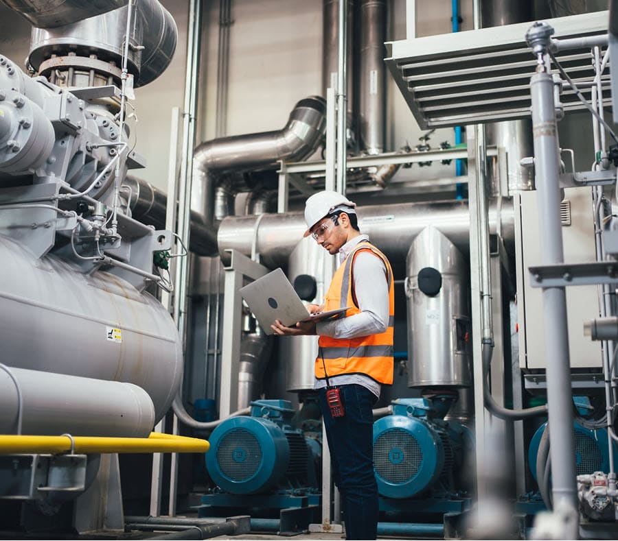 Engineer in a hard hat and safety vest using a laptop to monitor industrial equipment in a large facility, surrounded by machinery and pipes, performing real-time data analysis for system efficiency.